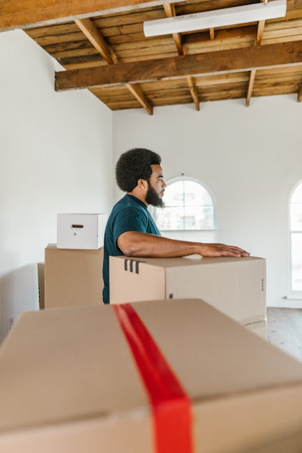 A man with curly hair and a beard, dressed in a blue shirt, stands inside a room with white walls and a wooden ceiling with exposed beams, during a home relocation process. He is positioned behind several cardboard boxes, one of which is in the foreground with a red packing strap across its top. The boxes vary in size and are stacked or arranged on the floor, ready for moving. Behind him, two arched windows allow natural light into the space, and the room appears unfurnished, indicating an ongoing packing and loading process. The scene illustrates furniture transport and packing activities typical of a house removal, with Clapham Junction Removals providing expert moving services to facilitate efficient and careful loading of belongings into a van outside the property.