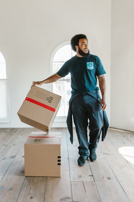 A man with curly hair and a beard, dressed in a blue shirt, stands inside a room with white walls and a wooden ceiling with exposed beams, during a home relocation process. He is positioned behind several cardboard boxes, one of which is in the foreground with a red packing strap across its top. The boxes vary in size and are stacked or arranged on the floor, ready for moving. Behind him, two arched windows allow natural light into the space, and the room appears unfurnished, indicating an ongoing packing and loading process. The scene illustrates furniture transport and packing activities typical of a house removal, with Clapham Junction Removals providing expert moving services to facilitate efficient and careful loading of belongings into a van outside the property.