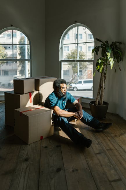 A man with curly dark hair and a beard, wearing a blue T-shirt and dark jeans, sits on the wooden floor inside a room with large, arched windows. The room is illuminated by natural daylight, showing a view of a street with parked cars and modern buildings outside. Behind him are several cardboard moving boxes, some sealed with red tape and others partially open. A tall potted plant with broad green leaves stands near the window. The scene depicts a moment during a home relocation or packing process, with the boxes ready for transport or unpacking, and the man appearing to rest between tasks. The setting and objects reflect professional moving and packing activities typically associated with house removals, as offered by Clapham Junction Removals.