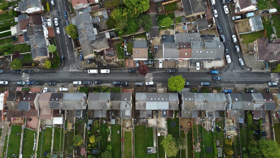 An aerial view of a residential street captured from above, showing a row of terraced houses with pitched roofs, some with skylights and chimneys. Vehicles, including small cars and vans, are parked along the curbside on both sides of the street. The street features a central lane without parking, with a few cars traveling along it. Behind the houses, garden yards are visible, some with trees, lawns, and outdoor furniture. The scene is well-lit with natural daylight, highlighting the details of the rooftops, street layout, and surrounding greenery. This overview image relates to house removals and moving services, illustrating the typical environment where such activities occur, particularly on narrow streets like Northcote Road in Clapham SW11 where space constraints influence the logistics of furniture transport and home relocation carried out by companies such as Clapham Junction Removals.