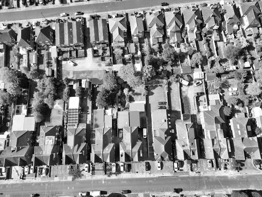 A black and white aerial photograph of a residential street showing a densely packed row of terraced houses with tiled roofs, narrow front gardens, and surrounded by mature trees. The street appears quiet with parked cars along the pavement. In the foreground, a large van is parked on the pavement, partially inside a driveway adjacent to a property, indicating a loading or unloading activity. Inside the van, visible packing materials such as cardboard boxes, packing paper, and a few pieces of furniture are being loaded or unloaded. Several individuals are engaged in carrying boxes and larger items, using equipment like trolleys and straps to assist with the furniture transport process. The scene captures a typical home relocation operation on a narrow street, with careful handling of household belongings by Clapham Junction Removals during a furniture transport and packing and moving activity in an urban residential environment.
